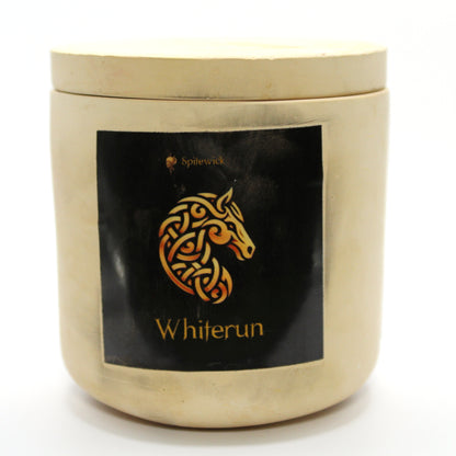 A beige candle holder with a sleek black label, featuring a horse intricately designed in stylized gold Celtic knotwork. Beneath the design, the name Whiterun is elegantly printed in gold, adding a bold yet sophisticated touch. Set against a clean white background, this handcrafted candle blends tradition with modern artistry, perfect for unique home decor, gifts, and personal expression.