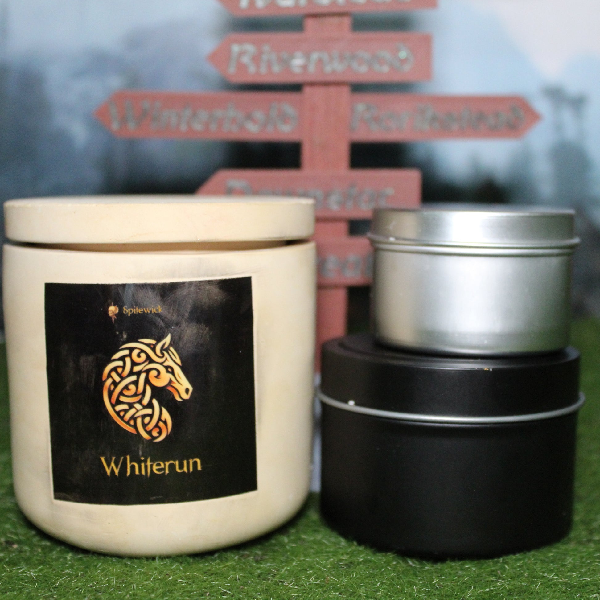 Front view of a handcrafted beige candle holder with a black label featuring a horse in gold Celtic knotwork, with the name whiterun in gold below. Two smaller candles—one black 4oz and one silver 2oz—show the three available sizes. Set on grass in front of a 3D-printed signpost marking Skyrim cities, blending medieval fantasy with artisan craftsmanship.