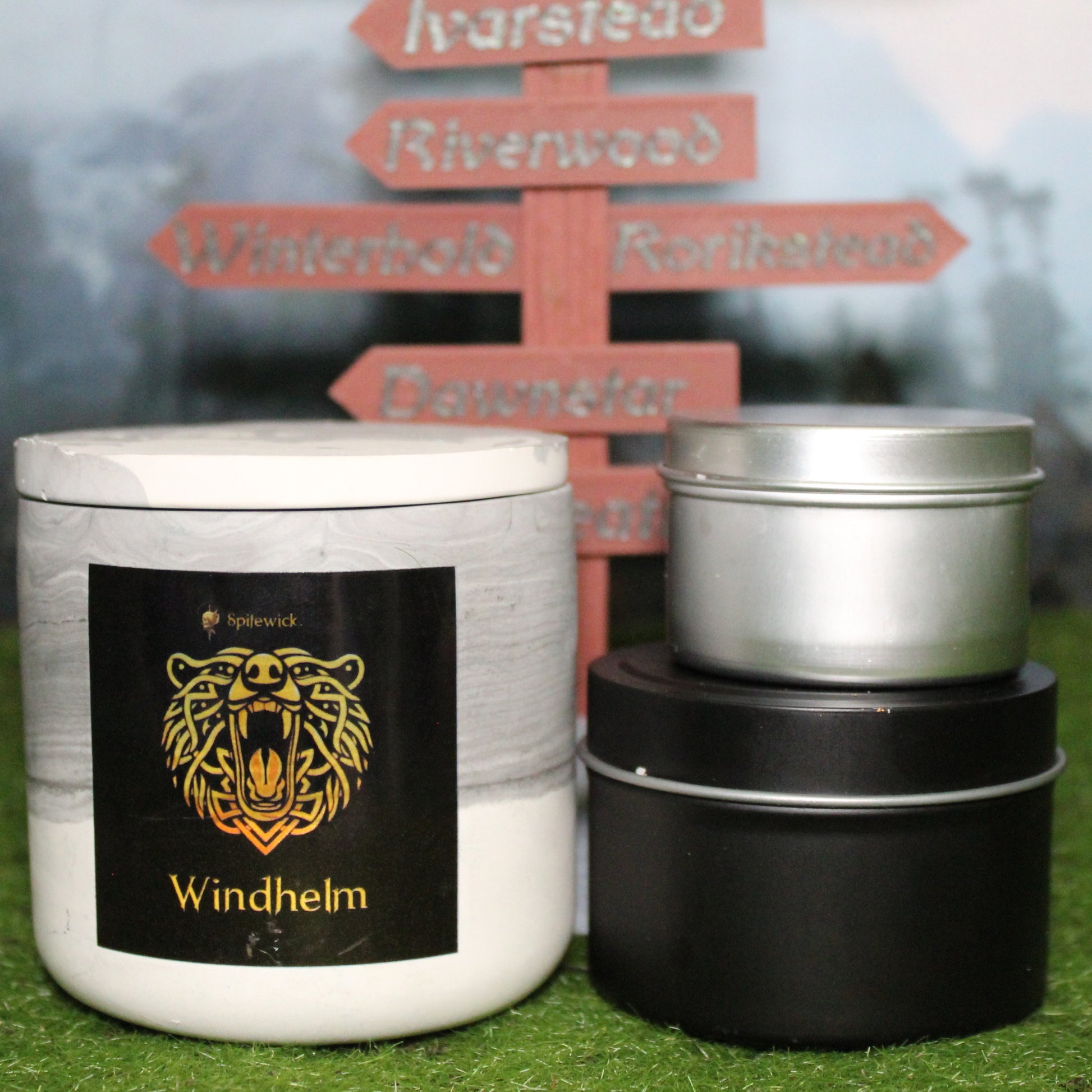 Front view of a handcrafted black and white candle holder with a black label featuring a bear in gold Celtic knotwork, with the name  windhelm in gold below. Two smaller candles—one black 4oz and one silver 2oz—show the three available sizes. Set on grass in front of a 3D-printed signpost marking Skyrim cities, blending medieval fantasy with artisan craftsmanship.