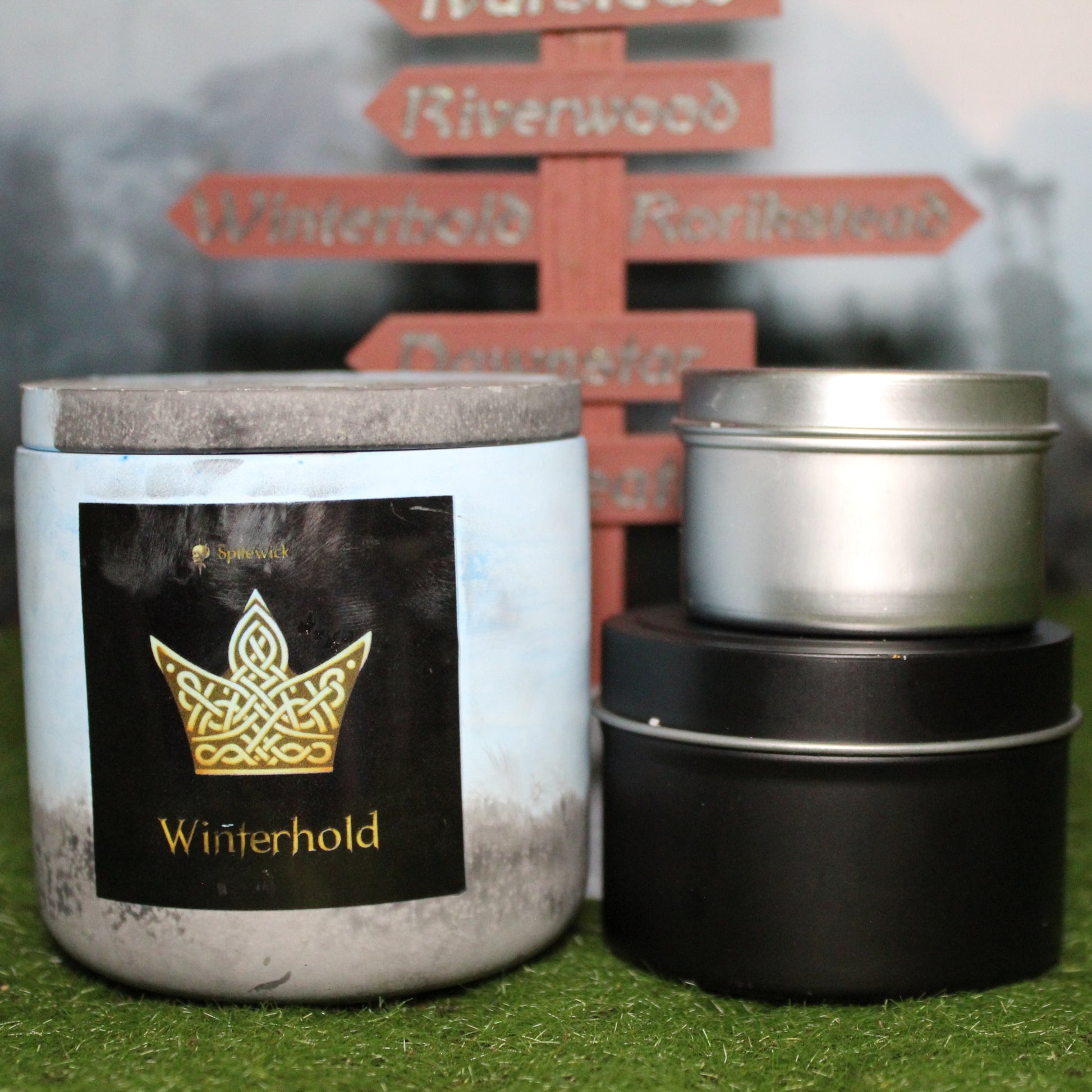 Front view of a handcrafted blue and black candle holder with a black label featuring a Crown in gold Celtic knotwork, with the name winterhold in gold below. Two smaller candles—one black 4oz and one silver 2oz—show the three available sizes. Set on grass in front of a 3D-printed signpost marking Skyrim cities, blending medieval fantasy with artisan craftsmanship.